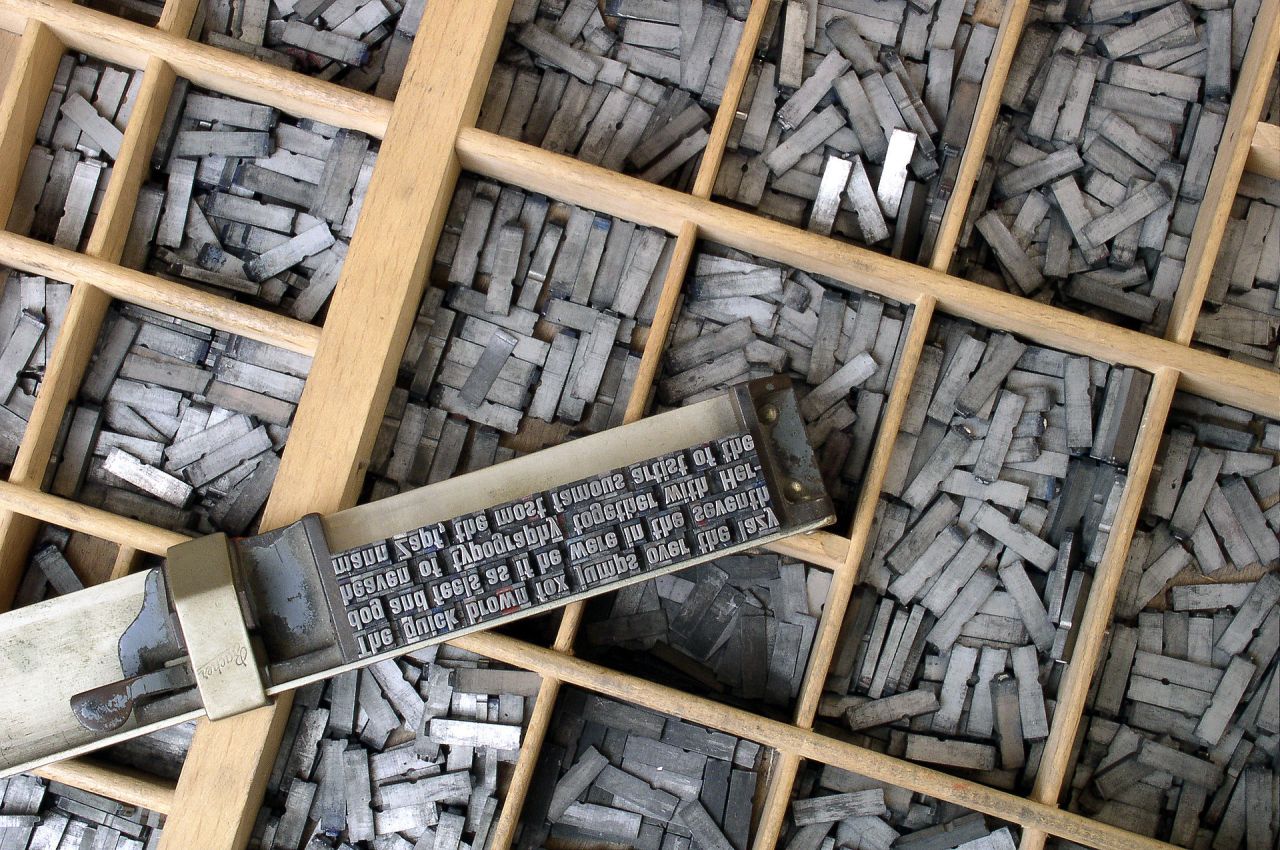 Moveable type on a composing stick in front of a type case. It reads, "The quick brown fox jumps over the lazy dog and feels as if he were in the seventh heaven of typography together with Hermann Zapf, the most famous artist of the"